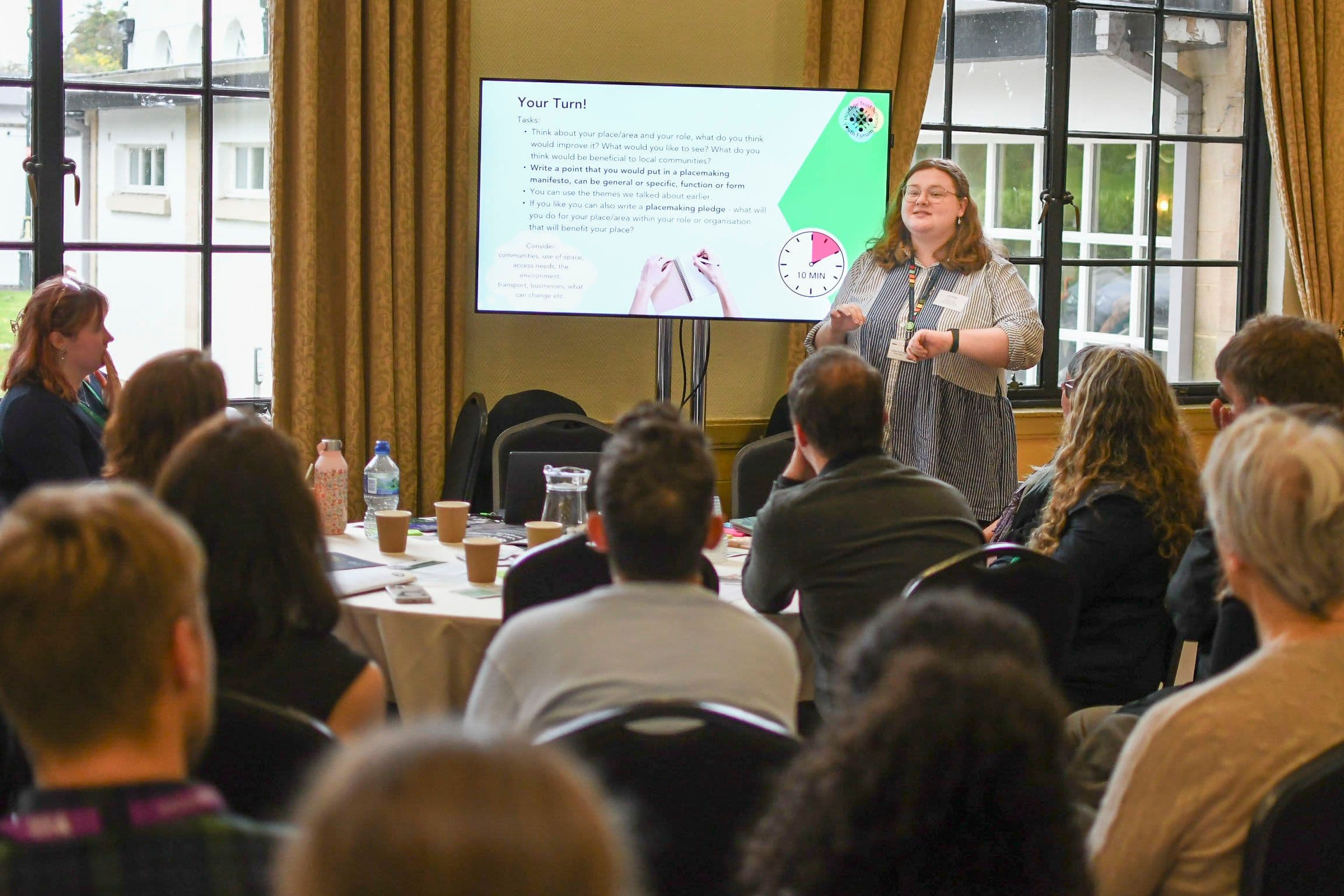 A group of people sit around looking at a woman who is giving a presentation on a screen.