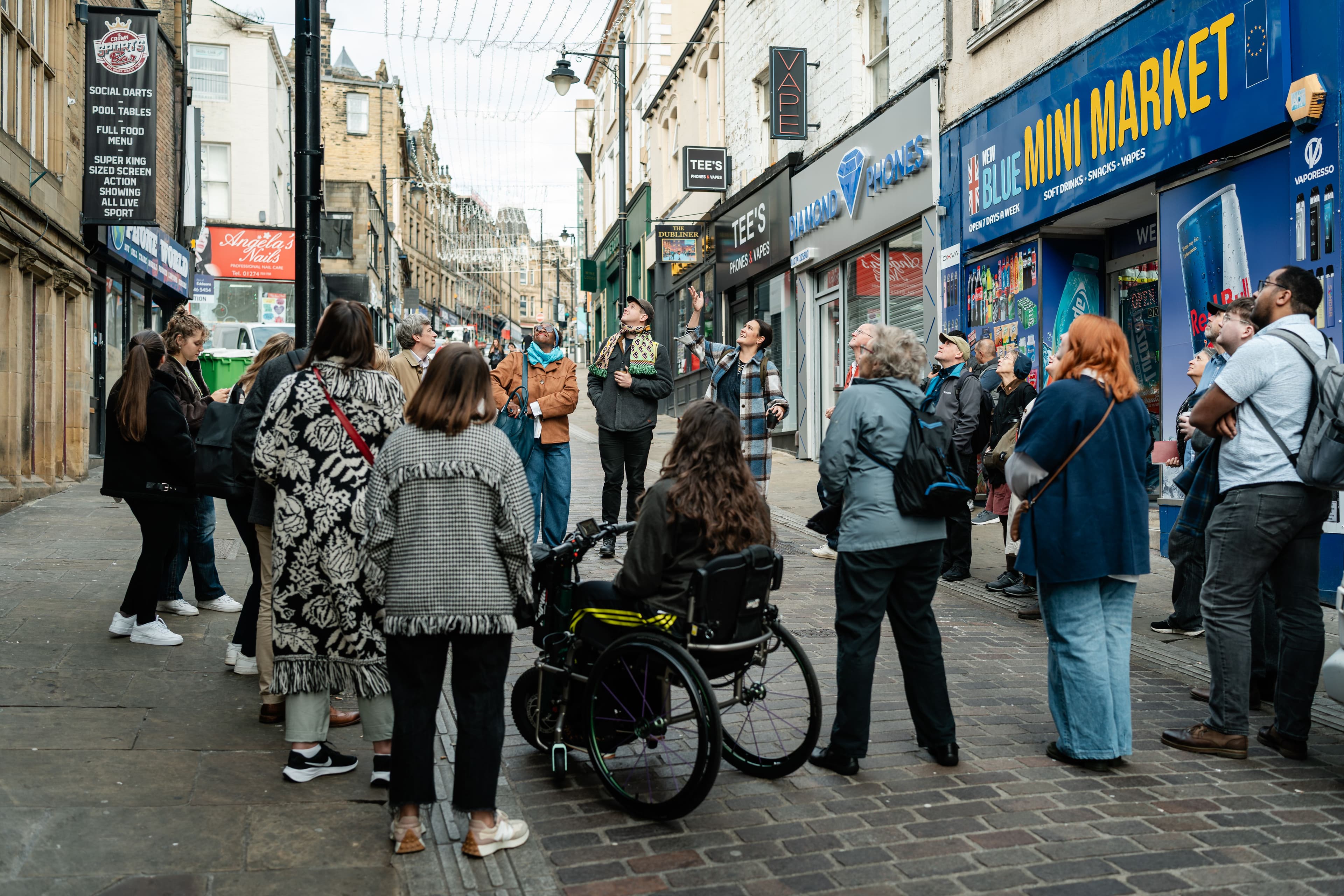 A photograph our a walking tour in Bradford. The group are gathered round the guide who is pointing out features on the buildings either side.