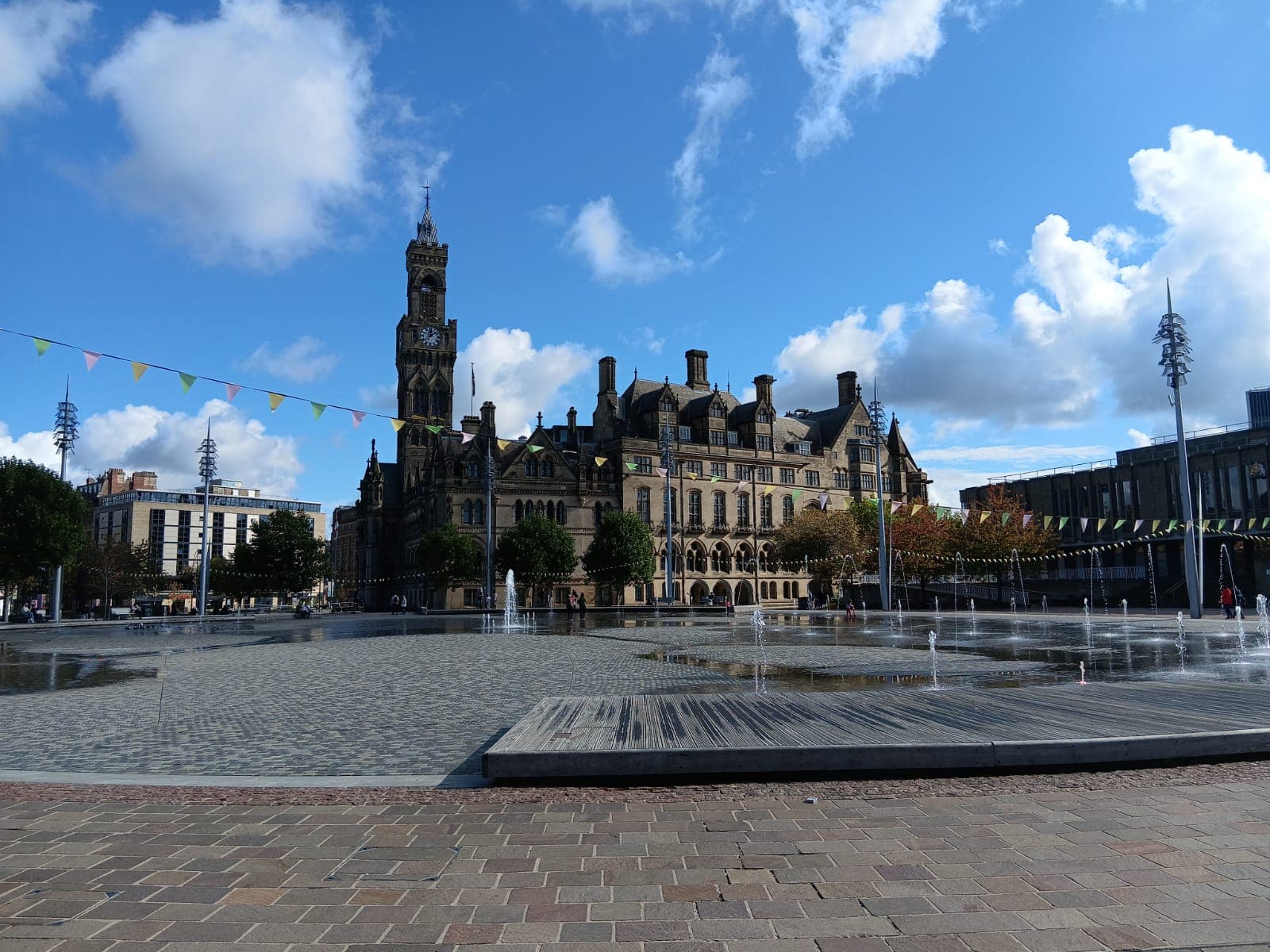A photograph looking across fountains towards Bradford Town Hall. It's bright and sunny and bunting hangs across the space.