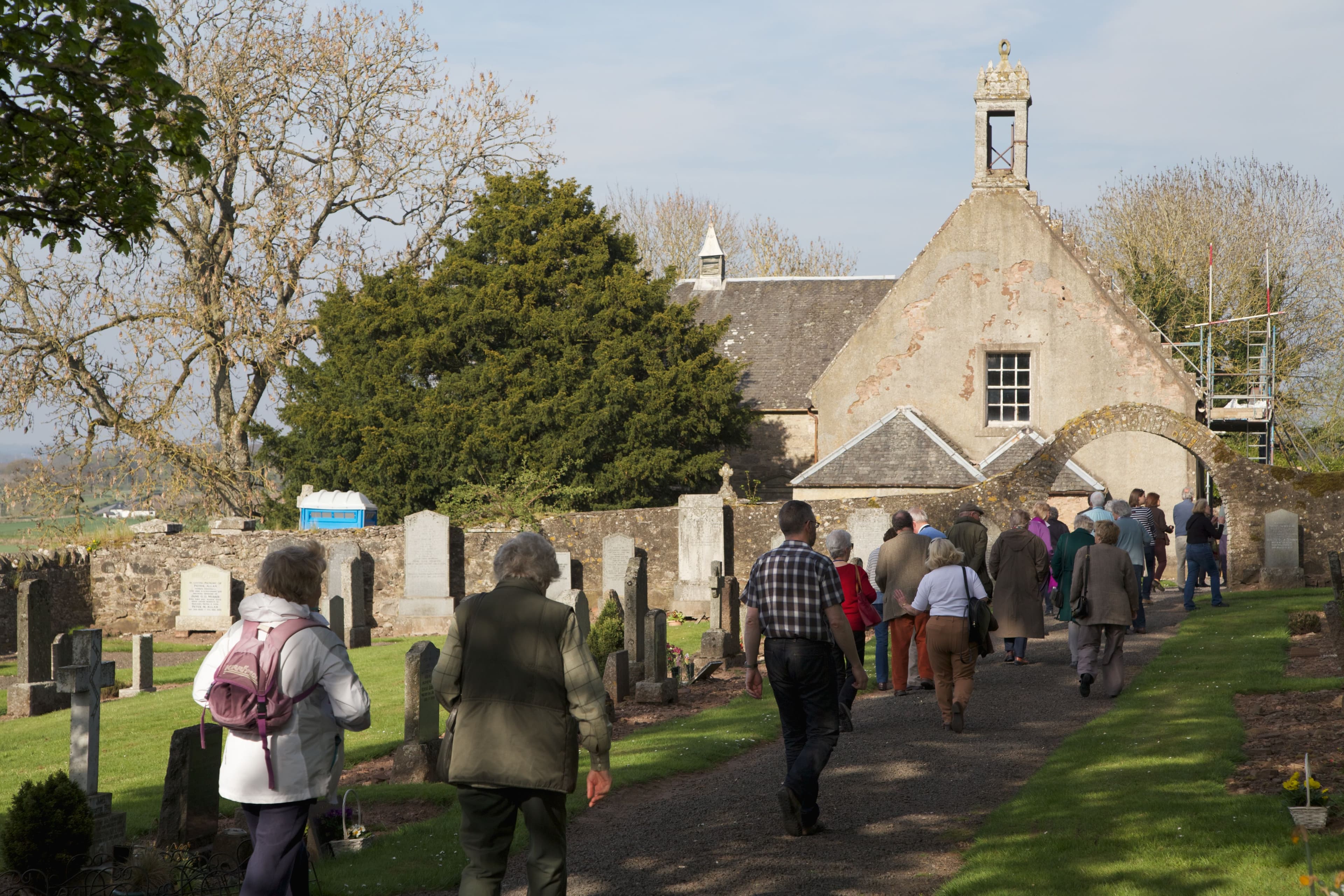 A path leads through a graveyard towards Tibbermore Parish Church. A string of people walk down the path towards the building which has scaffolding up the side.